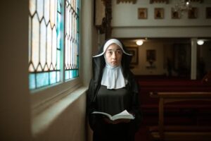 A nun is standing inside a church by the stained glass, holding a Bible.