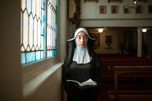 A nun is standing inside a church by the stained glass, holding a Bible.